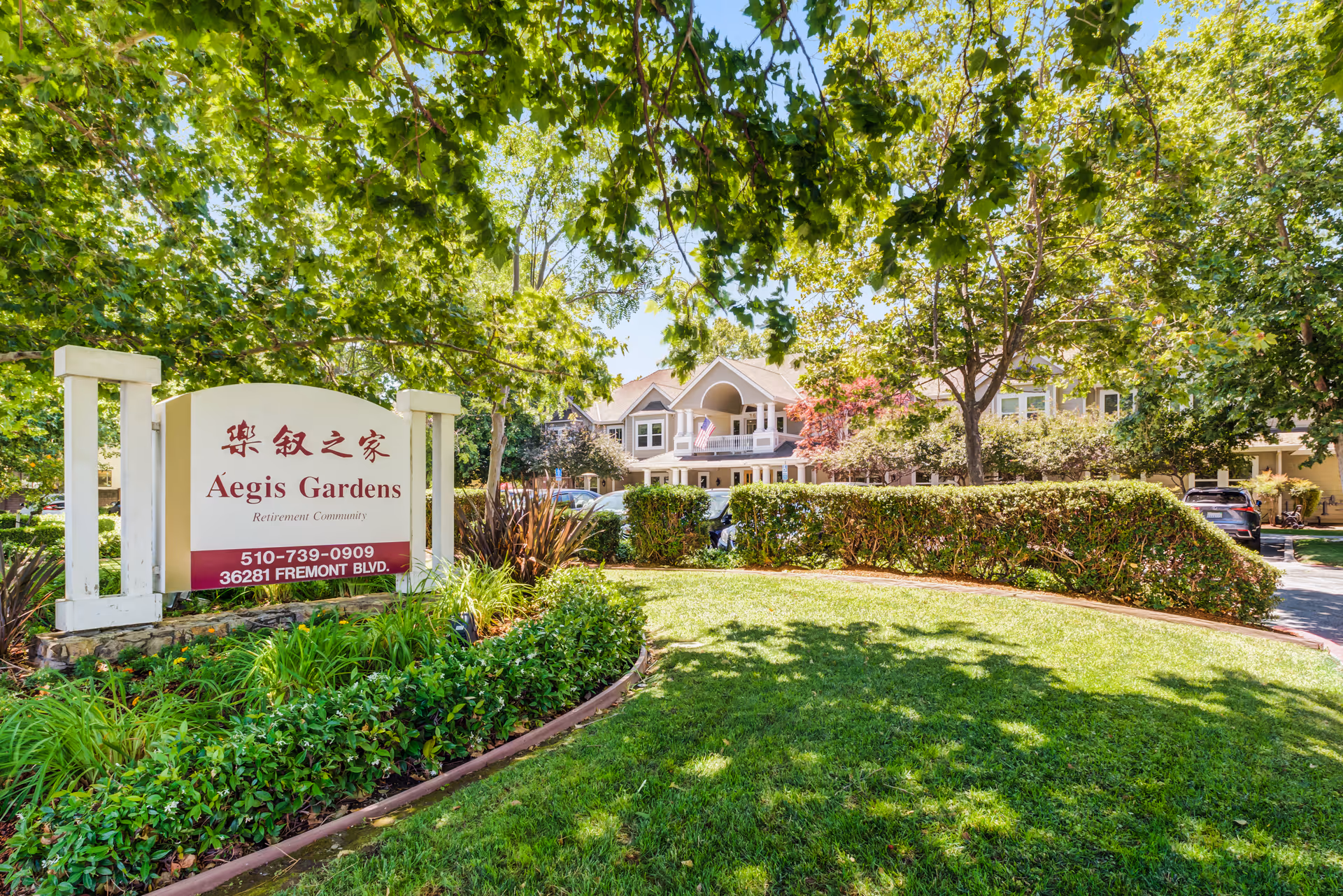 Aegis Gardens Fremont retirement community sign surrounded by green bushes and trees with a large residential building and parked cars in the background under a clear blue sky.