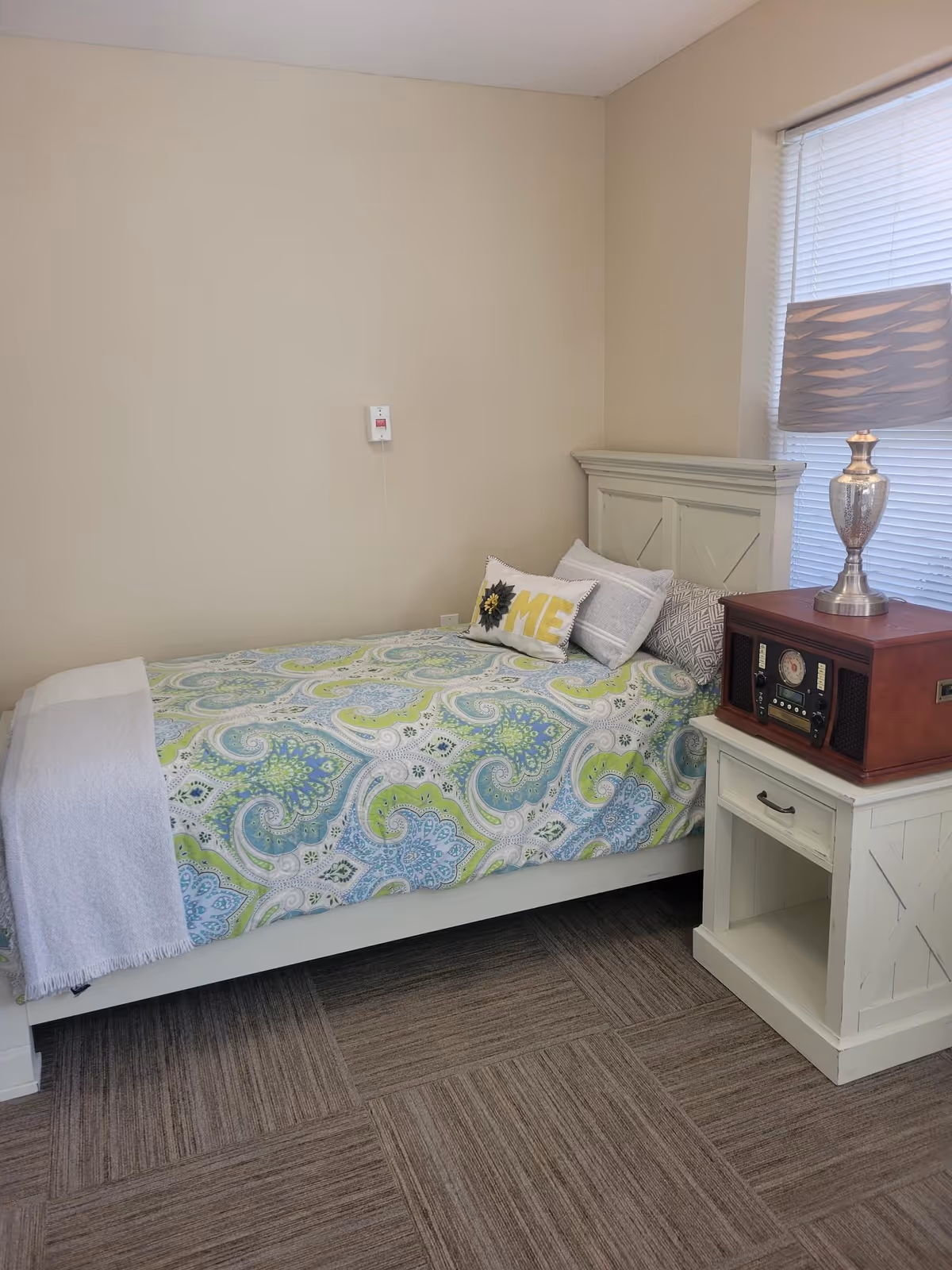A small single bedroom with a bed dressed in a green-and-blue paisley comforter, a white headboard, and a nightstand holding a lamp and vintage-style radio.