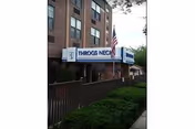 Exterior view of Throgs Neck Rehabilitation & Nursing Center showing a multi-story brick building with a white and blue sign displaying the facility's name. There is an American flag on a pole near the entrance and green bushes along a fenced walkway.