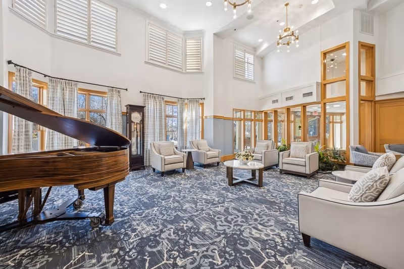 A spacious and bright living room area with a grand piano on the left, several cushioned armchairs arranged around a round coffee table, large windows with curtains, a tall grandfather clock, and high ceilings with chandeliers.