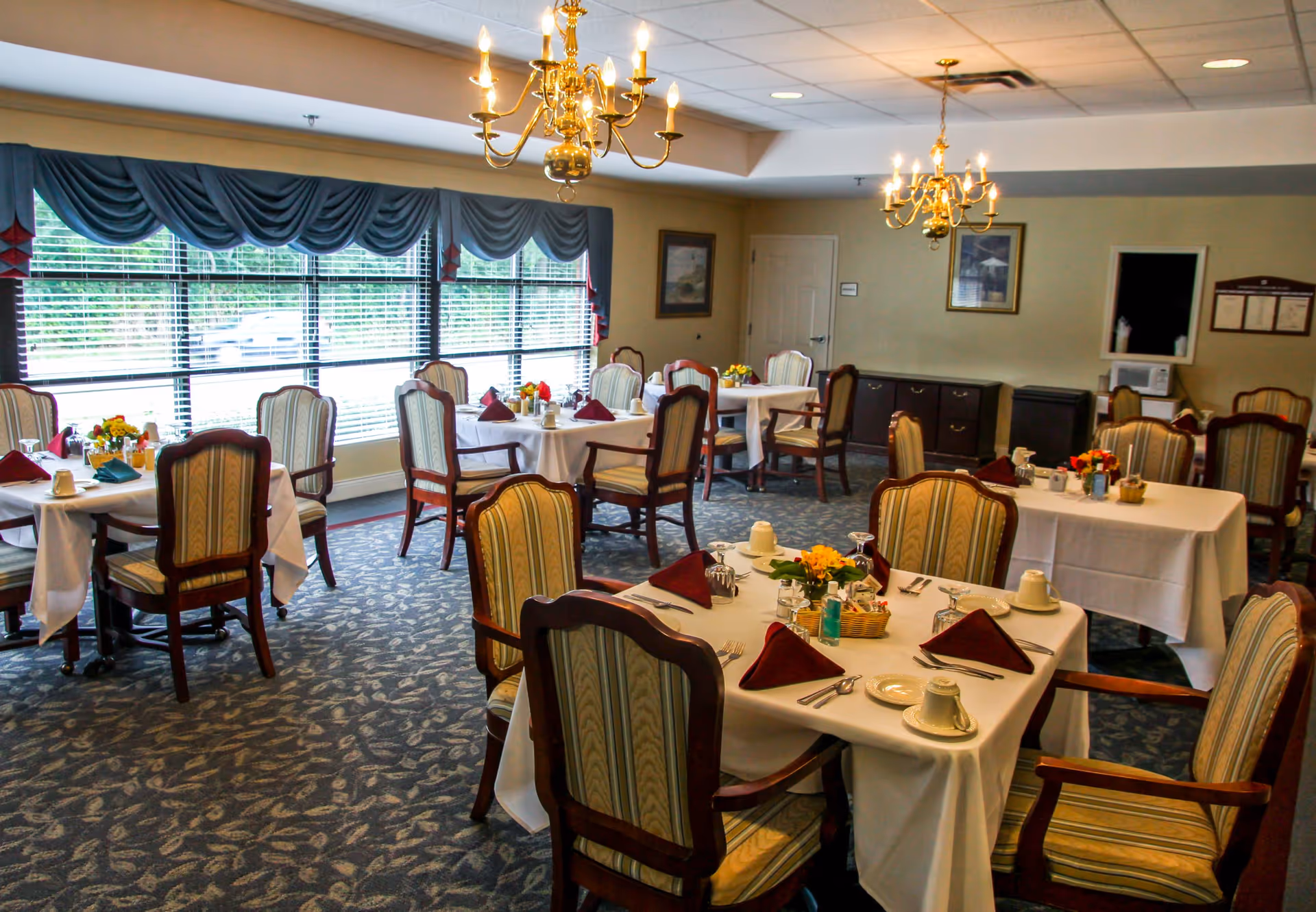 Well-lit dining room with multiple set tables, upholstered chairs, chandeliers, and large windows.