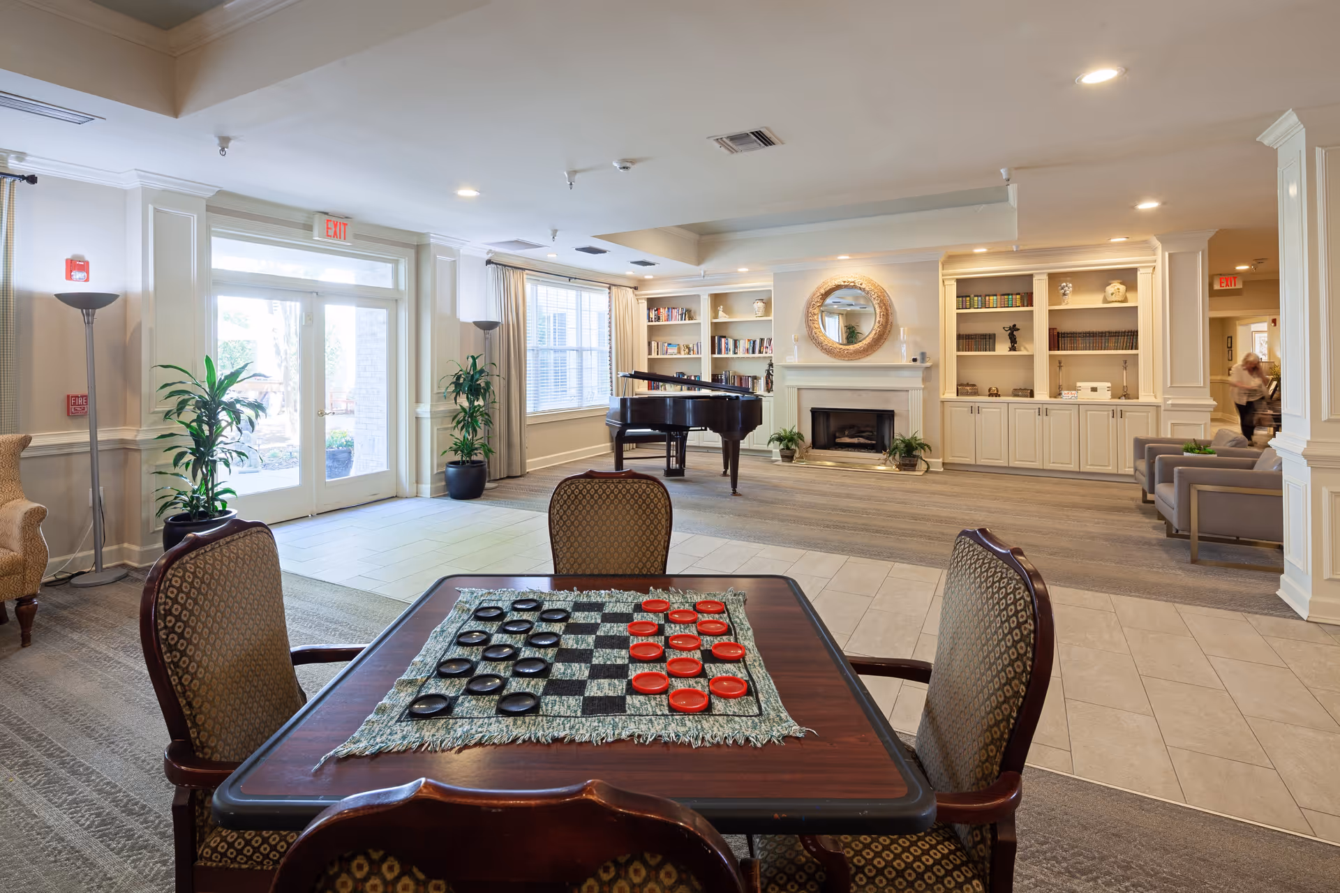 A spacious and well-lit common area in a senior living facility featuring a table with a checkers game set up, comfortable chairs, a grand piano, built-in bookshelves, a fireplace with a round mirror above it, potted plants, and large windows letting in natural light.