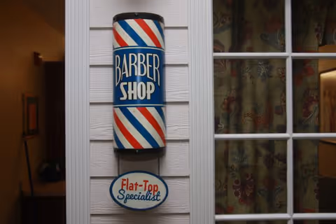 A traditional barber shop pole mounted on a white wall next to a window with floral curtains. The pole has red, white, and blue stripes with the words 'BARBER SHOP' and a smaller sign below that reads 'Flat-Top Specialist'.
