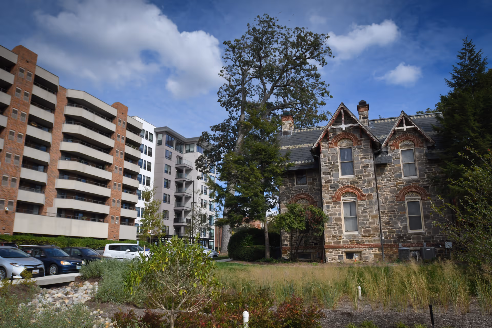 Exterior view of Roland Park Place showing a modern multi-story apartment building on the left and an older stone house with brick accents on the right, surrounded by trees, grass, and a parking area with cars under a partly cloudy sky.