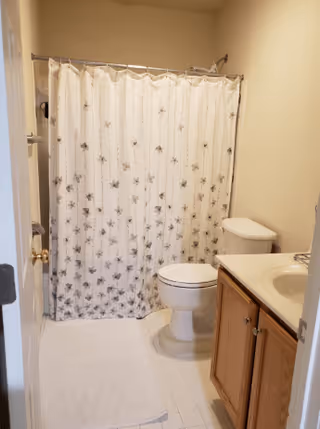 A small bathroom with a white shower curtain featuring a floral pattern, a white toilet, a wooden vanity with a white sink, and a white bath mat on the floor.