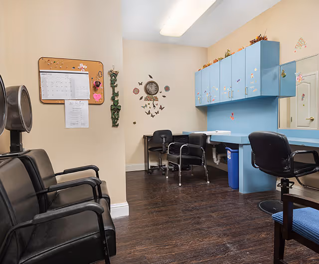 Small salon-style room with black styling chairs, a counter with mirrors, blue wall cabinets, and decorative wall clock and butterflies.