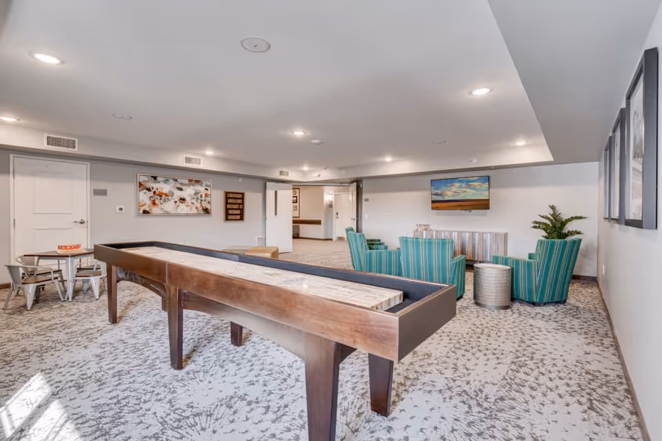 A spacious common room with a shuffleboard table in the foreground, a small table with chairs to the left, and a seating area with four striped green armchairs and a round metallic side table facing a wall-mounted TV. The room has carpeted floors, light gray walls, recessed ceiling lights, and framed artwork on the walls.