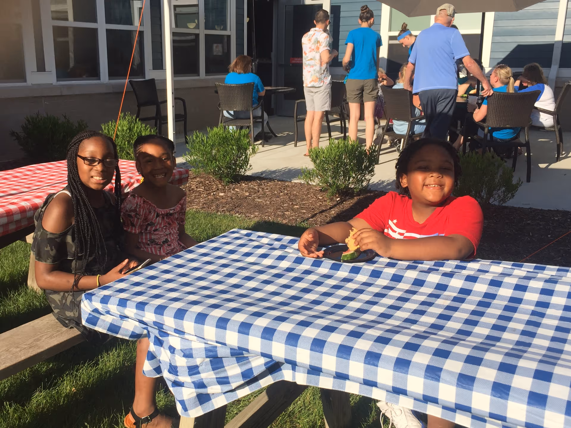 Three children sit at a picnic table with a blue-and-white checkered tablecloth in an outdoor courtyard, with adults and chairs near a building in the background.