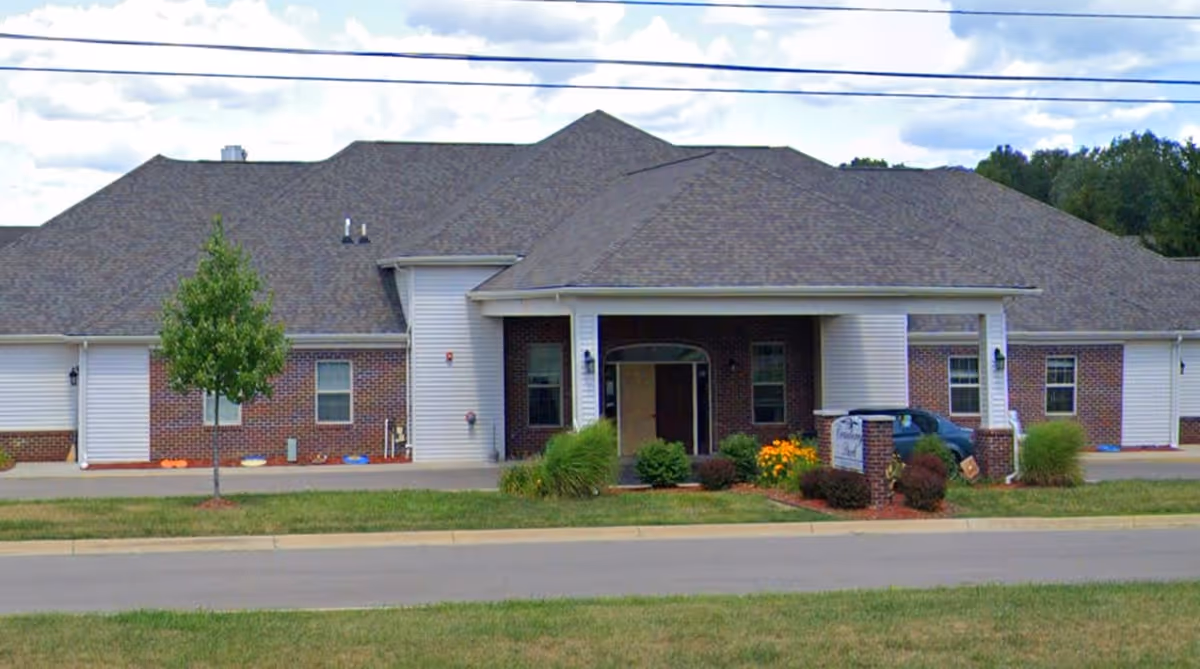 Front exterior view of a single-story brick and siding building with a covered entrance, a small tree, shrubs, and a sign in front. The sky is partly cloudy.