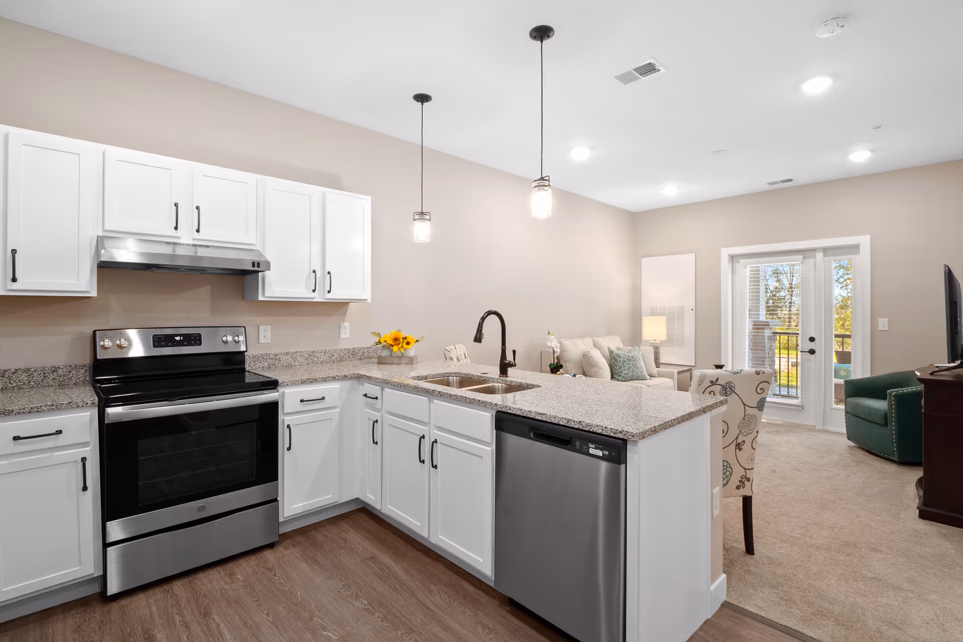 Modern kitchen with white cabinets, granite countertops, stainless steel appliances including an oven and dishwasher, and two pendant lights hanging above the counter. The kitchen opens into a living area with a beige sofa, patterned chair, green armchair, and a TV on a wooden stand. There are glass doors leading to an outdoor space.