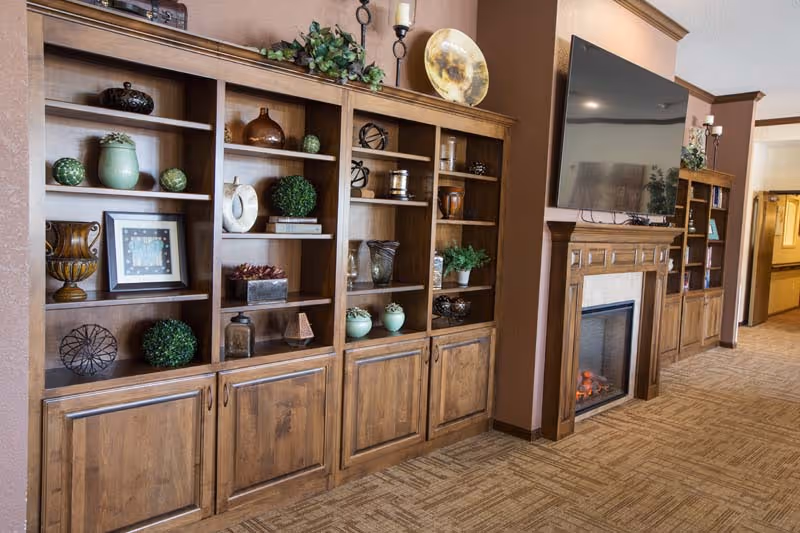 A cozy interior hallway featuring built-in wooden shelves decorated with various vases, plants, and decorative items. There is a fireplace with a mounted flat-screen TV above it, and the carpeted floor extends down the hallway with warm-toned walls.