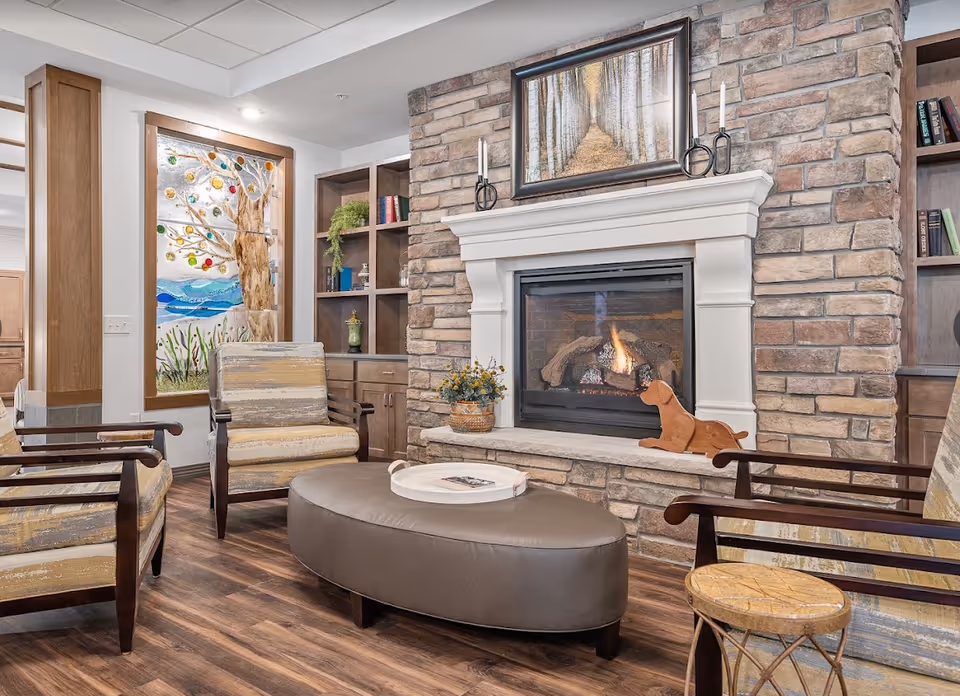 Cozy sitting area with upholstered armchairs and an ottoman arranged around a stone fireplace in a common living room.