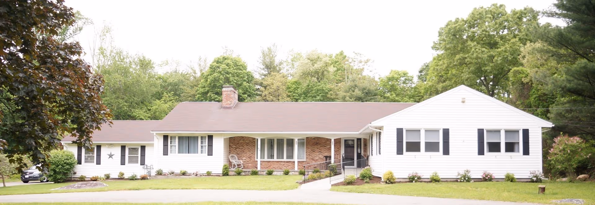 Single-story residential building with white siding and a brown roof, featuring a front porch with a chair and a ramp leading to the entrance. The house is surrounded by green grass, bushes, and trees in the background.