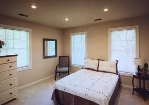 A bedroom with a double bed covered in a white and brown quilt, two large windows with blinds, a wooden chair, a white dresser with drawers, a wall mirror, and a small round side table with a lamp and a plant.
