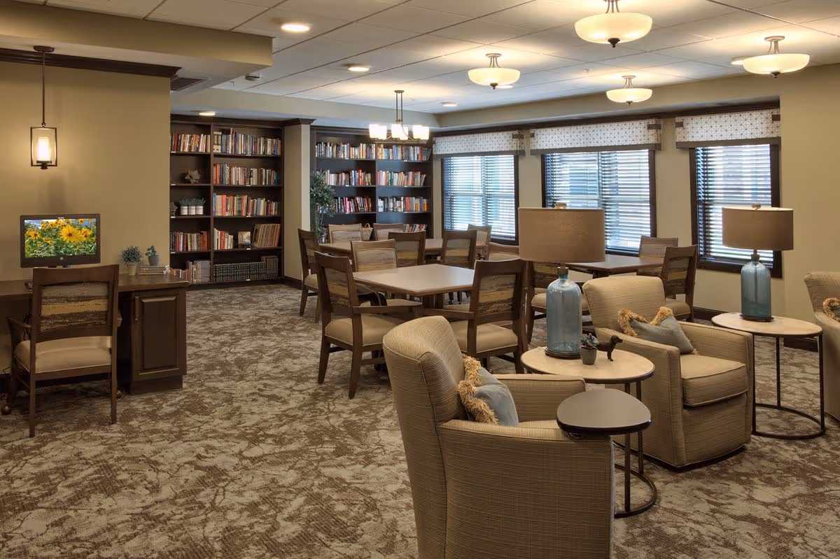 A cozy and well-lit common area in a senior living facility featuring comfortable armchairs with side tables and lamps, several square tables with wooden chairs, and a bookshelf filled with books along the back wall. There is a small desk with a computer displaying a sunflower image on the screen. Large windows with blinds allow natural light to enter the room.