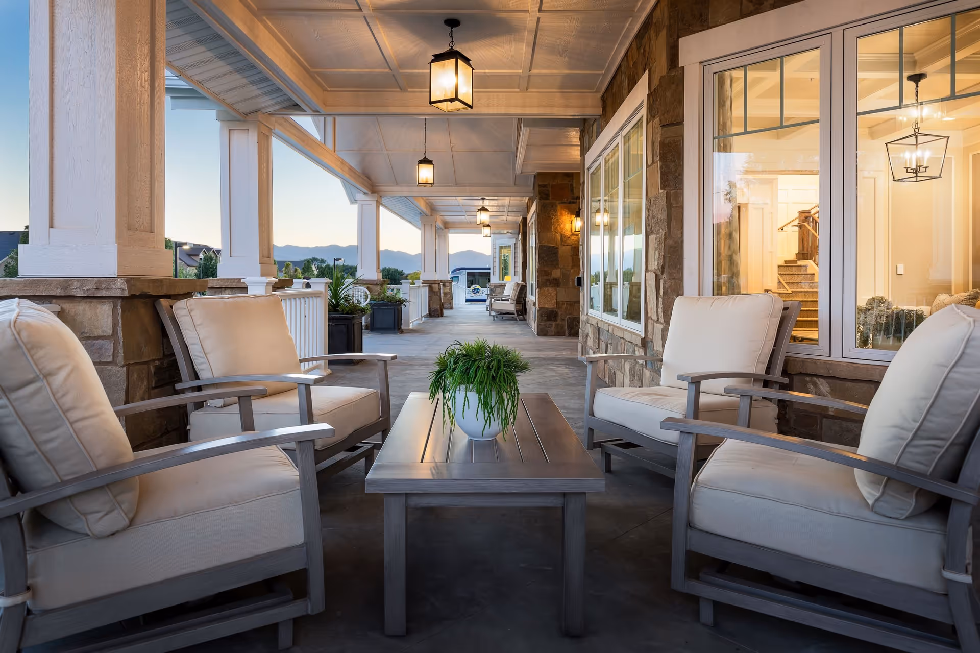 Covered outdoor patio area with cushioned armchairs arranged around a wooden coffee table with a potted plant. The patio features stone pillars, hanging lantern-style lights, and large windows showing an interior staircase and chandelier. Mountains and a clear sky are visible in the background.