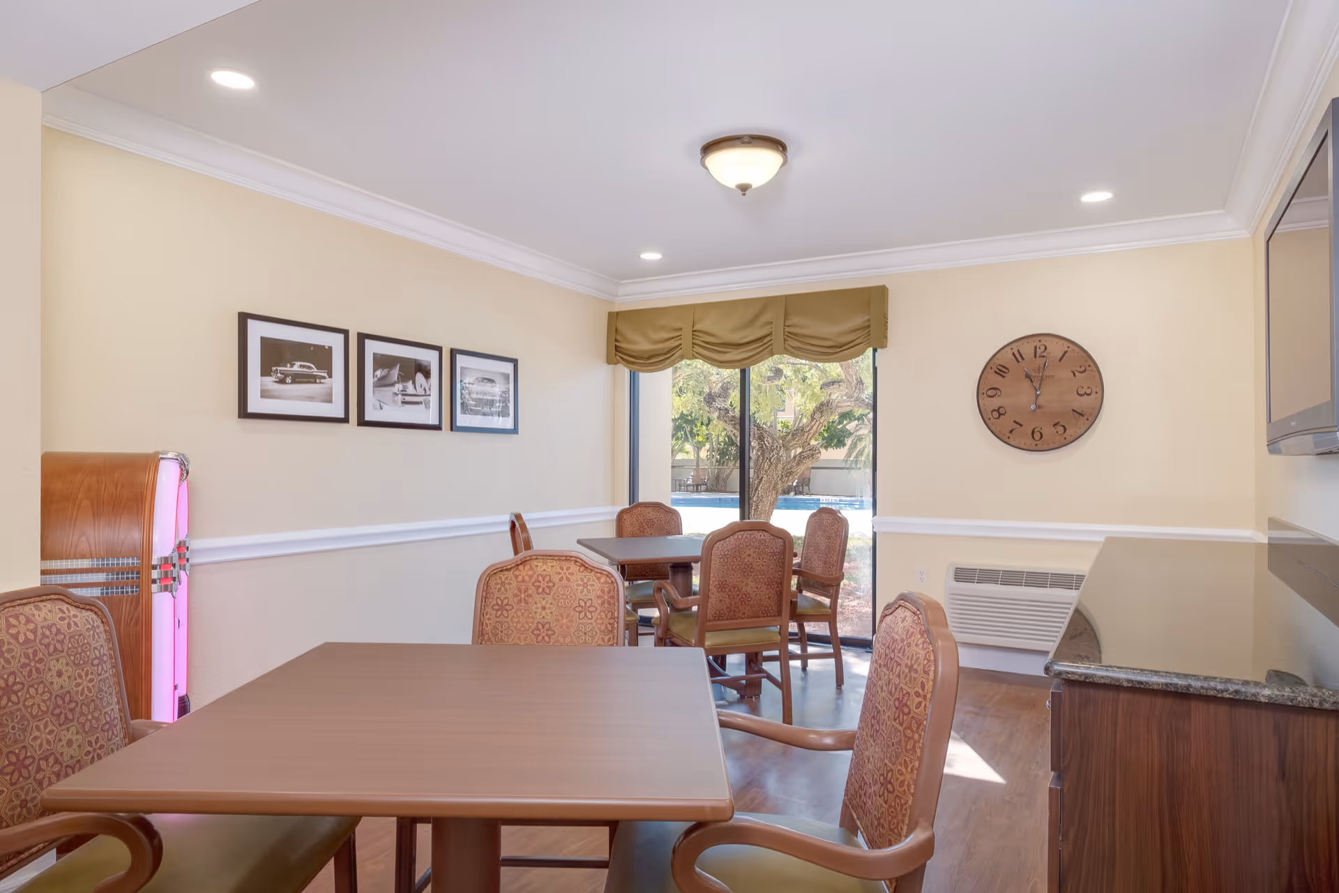 A bright dining room with wooden tables and cushioned chairs featuring floral upholstery. The room has beige walls with white trim, three black and white framed photographs on one wall, a large round clock on another wall, and a window with a green valance showing a tree and outdoor area. There is a jukebox with pink lighting in the corner and a wooden cabinet with a granite countertop on the right side.