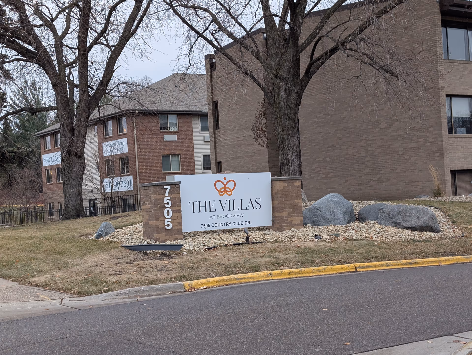 Outdoor view of The Villas at Brookview senior living facility sign with address 7505 Country Club Dr. The sign is placed on a landscaped area with rocks and grass, in front of a multi-story brick building and leafless trees.