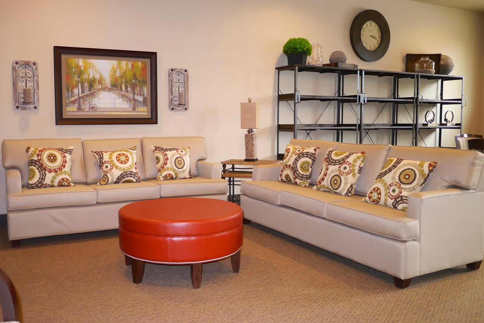 A cozy living room with two beige sofas adorned with patterned throw pillows, a round red ottoman in the center, a wooden side table with a lamp, a black metal shelving unit with decorative items, a large wall clock, and a framed painting of a canal scene on the wall.