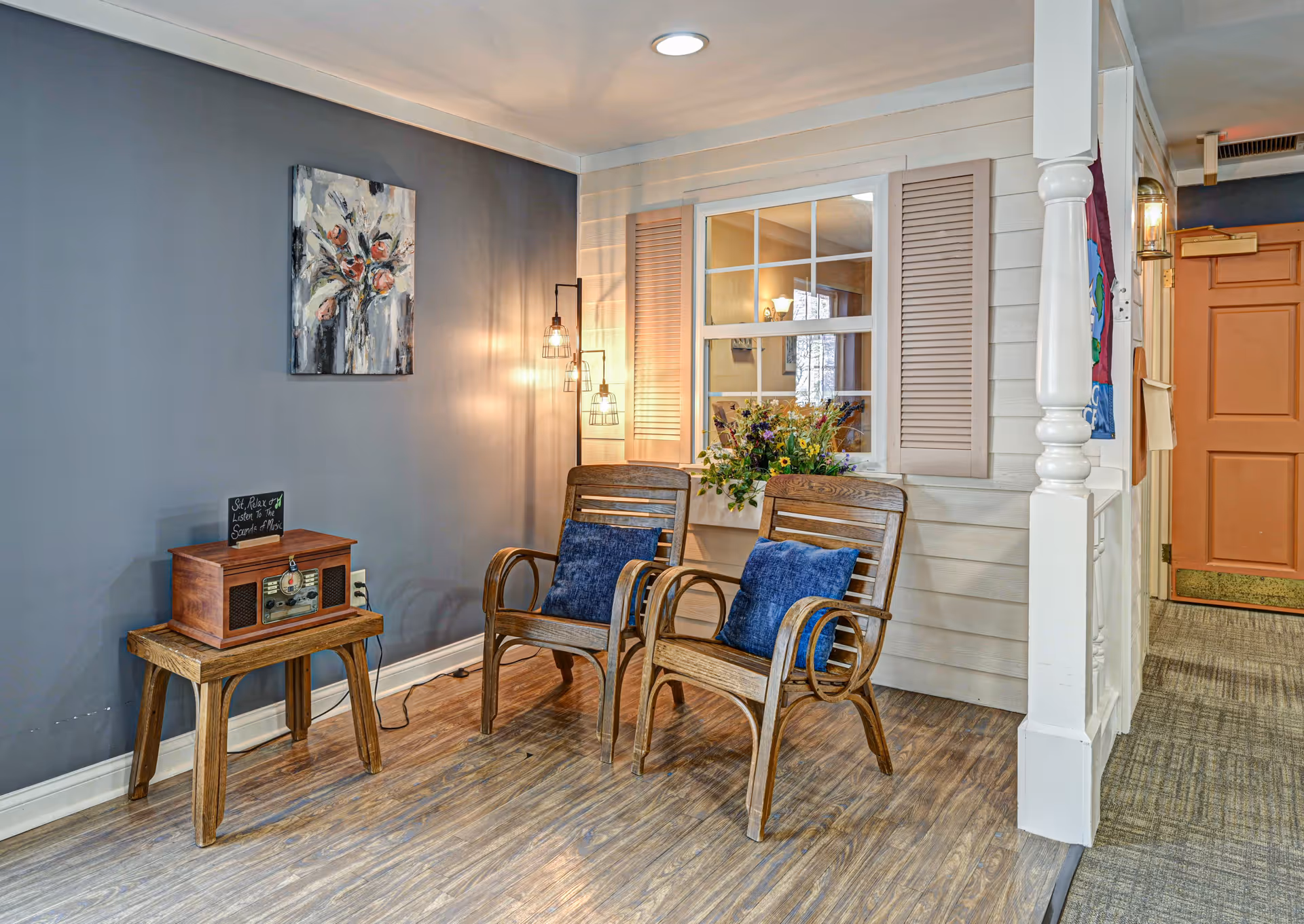 A cozy seating area with two wooden chairs featuring blue cushions, a small wooden table with a vintage-style radio, and a wall-mounted painting of flowers. The wall behind the chairs is painted gray, and there is a window with beige shutters and a flower arrangement on the windowsill. Warm lighting fixtures hang on the wall, and a wooden door is visible in the background.
