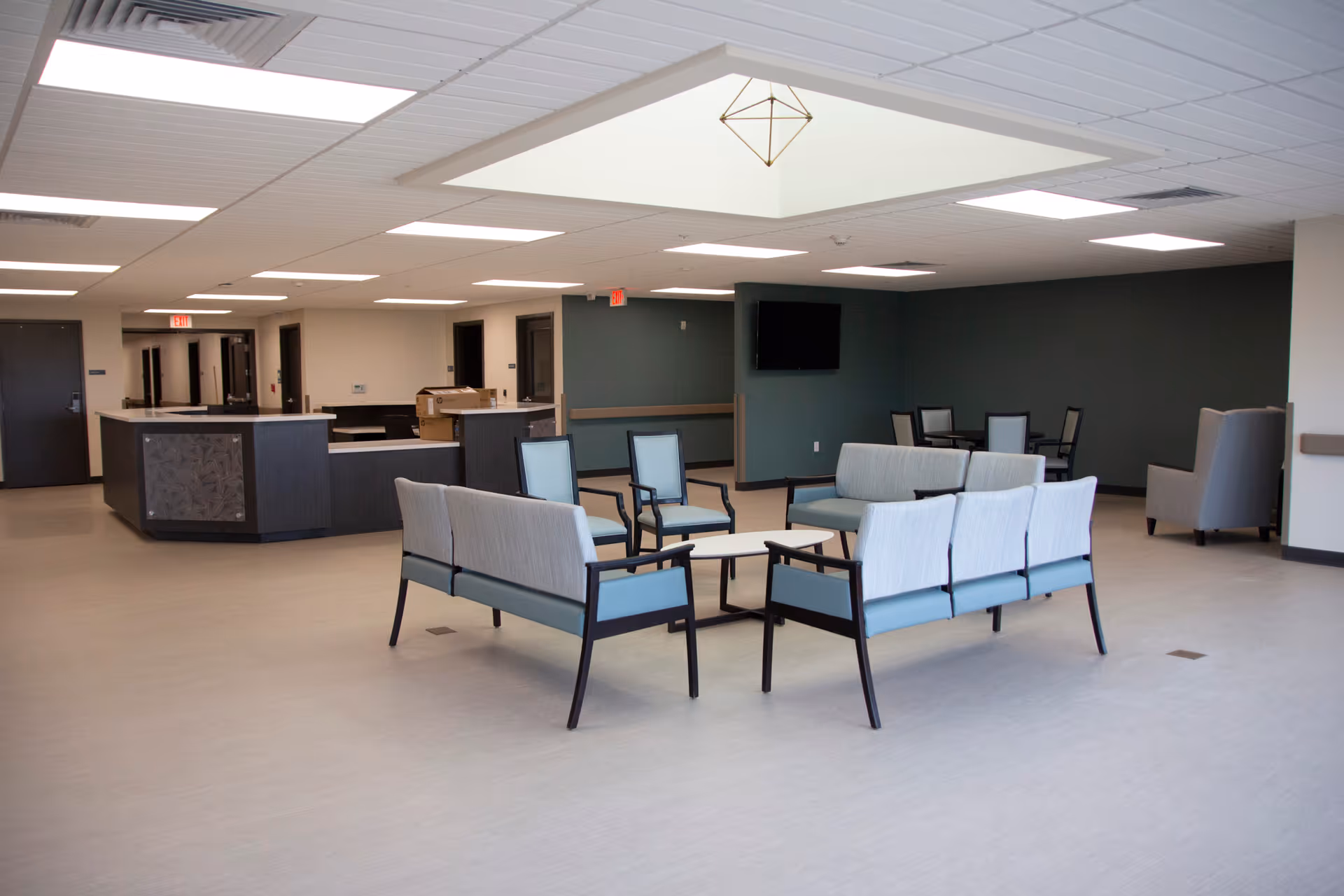 A spacious and modern common area in a senior living facility with multiple seating arrangements including sofas and chairs around small tables. The room has a light-colored floor, a dark accent wall with a mounted flat-screen TV, and a reception desk area in the background. The ceiling features recessed lighting and a skylight.