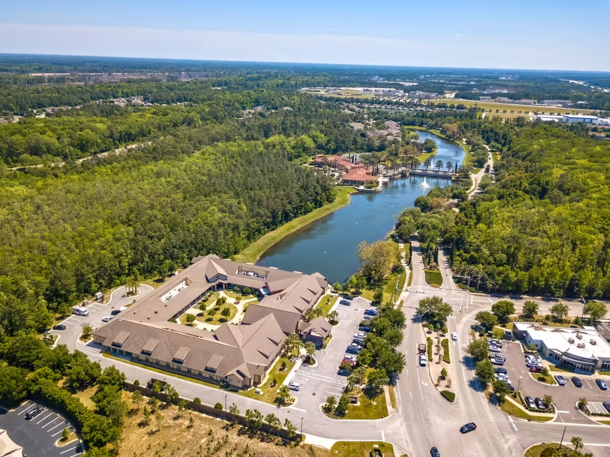 Aerial view of Rosecastle at Deerwood facility surrounded by lush green trees and a winding river. The building has a large parking lot with several cars parked. Roads and landscaped areas are visible around the facility under a clear blue sky.
