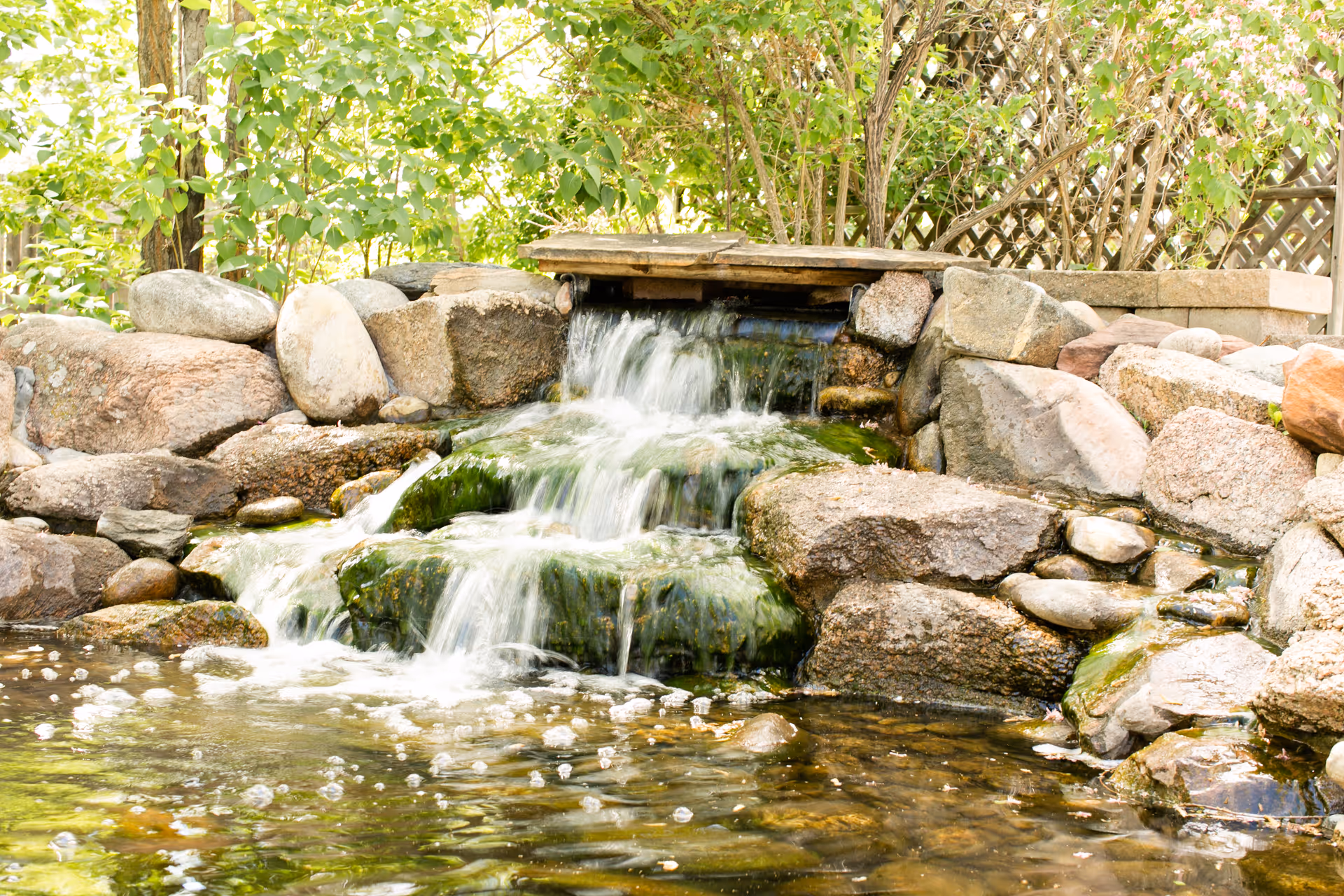 A small outdoor waterfall feature with water cascading over moss-covered rocks into a pond, surrounded by large stones and green leafy plants in the background.