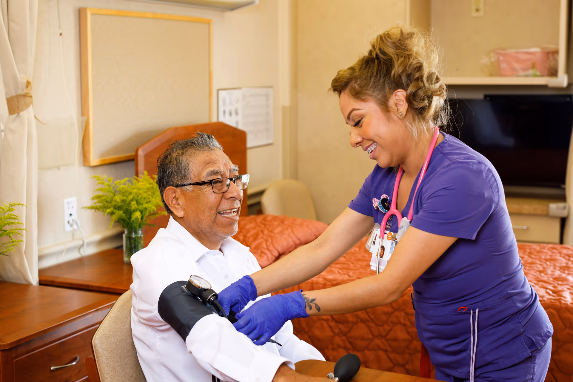 A healthcare worker in purple scrubs and blue gloves is smiling while taking the blood pressure of an elderly man wearing glasses and a white shirt. They are in a room with a bed covered in an orange quilt, a wooden dresser, and a vase with green plants.