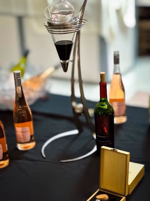 A table covered with a black cloth displaying several bottles of wine, including rosé and red wine. There is a modern wine decanter with a funnel-like design holding red wine above the table, and an open wooden box with a cork and corkscrew inside.