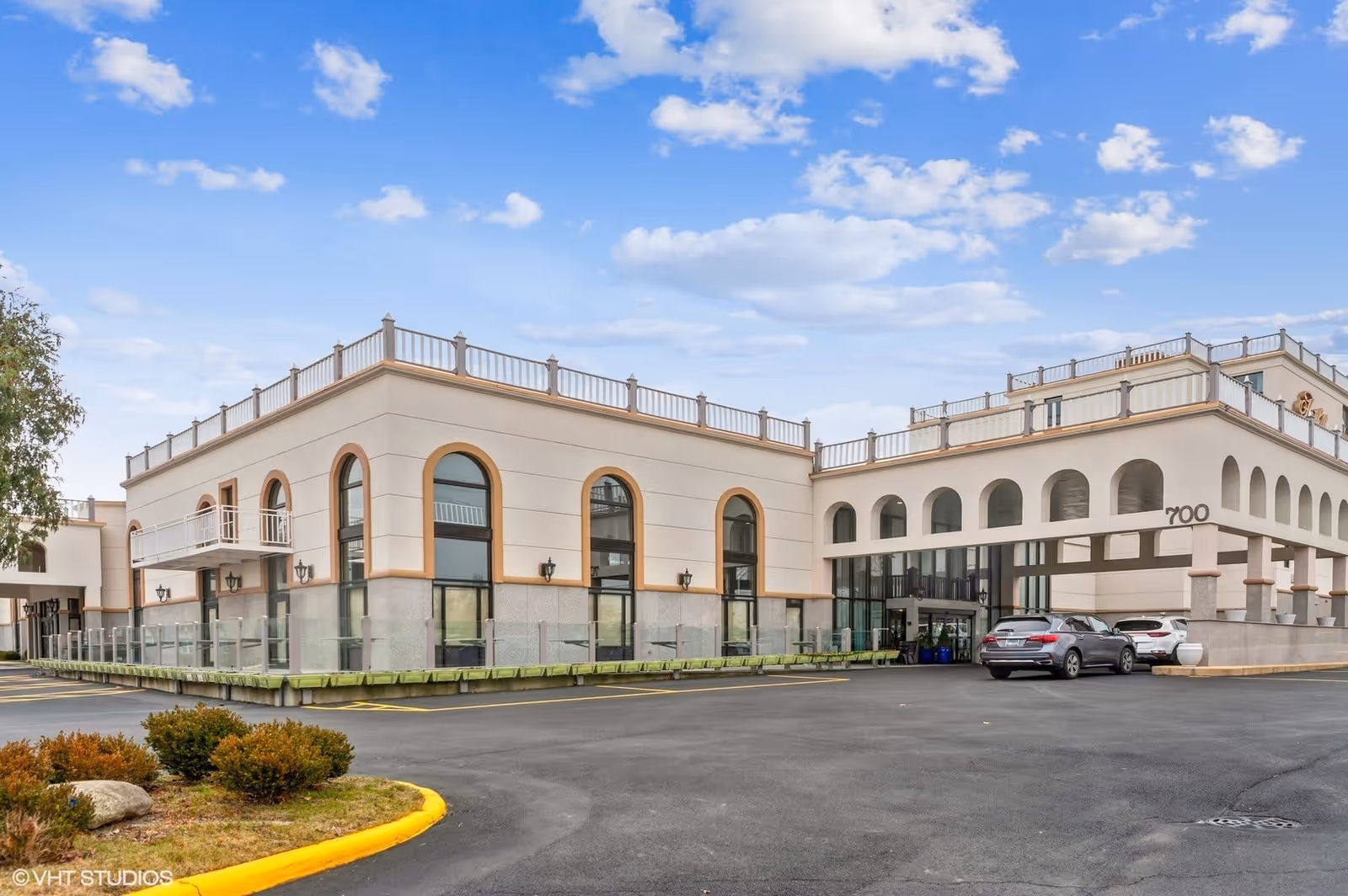 Exterior view of a large, light-colored building with arched windows and a covered entrance. There are two cars parked near the entrance, and the building has the number 700 displayed on the front. The sky is blue with scattered clouds.