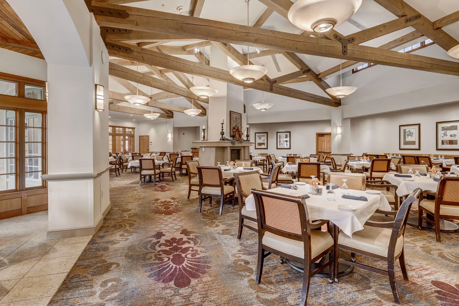Spacious dining room with multiple set tables, exposed wooden ceiling beams, and patterned carpet.