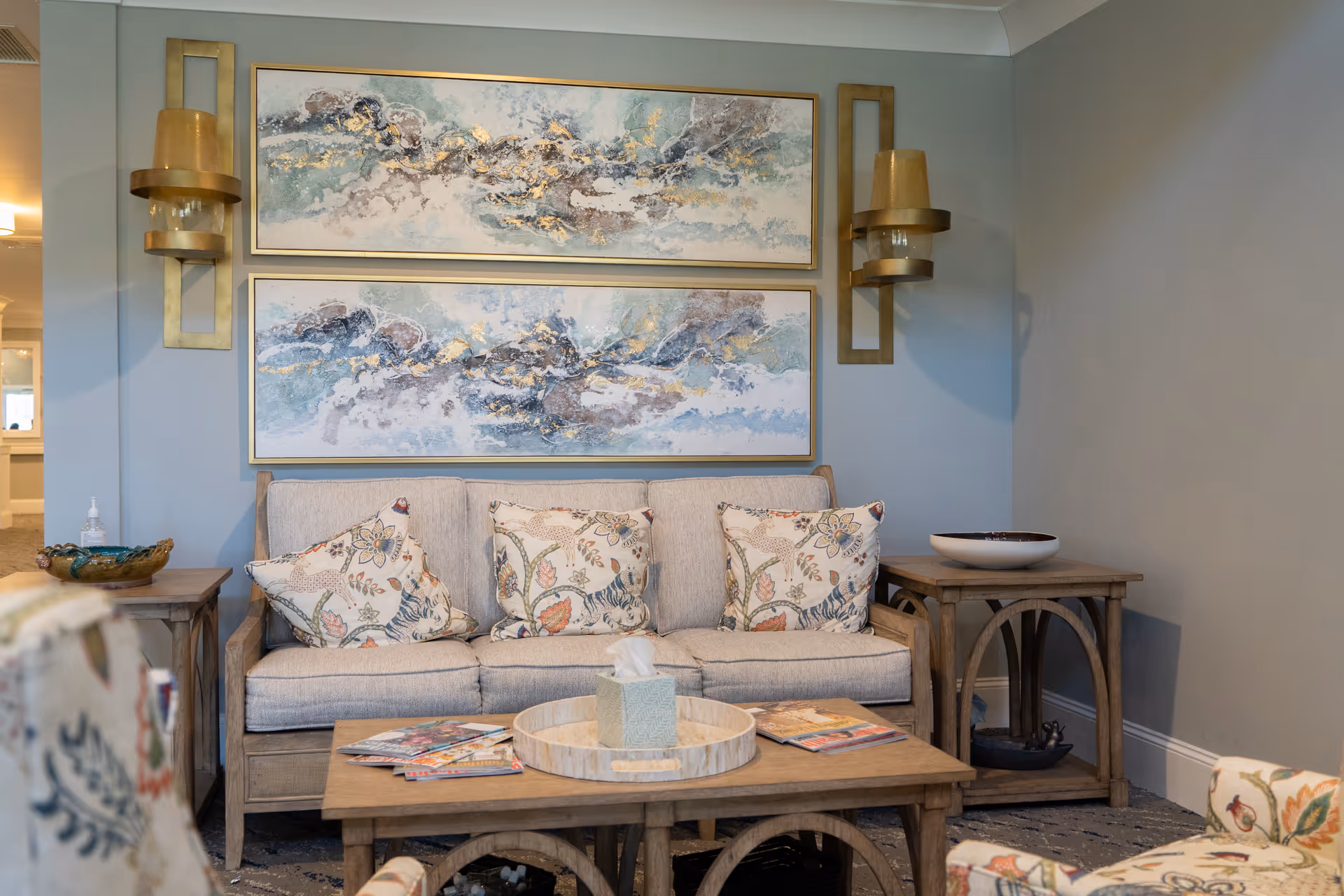 A cozy living room area with a beige sofa adorned with floral patterned pillows. Above the sofa are two abstract paintings with blue, white, and gold tones. On either side of the paintings are gold wall sconces. In front of the sofa is a wooden coffee table with magazines, a tissue box, and a decorative tray. Matching wooden side tables flank the sofa, one with a decorative bowl and the other with a small sculpture underneath. The room has soft lighting and a calm, neutral color palette.