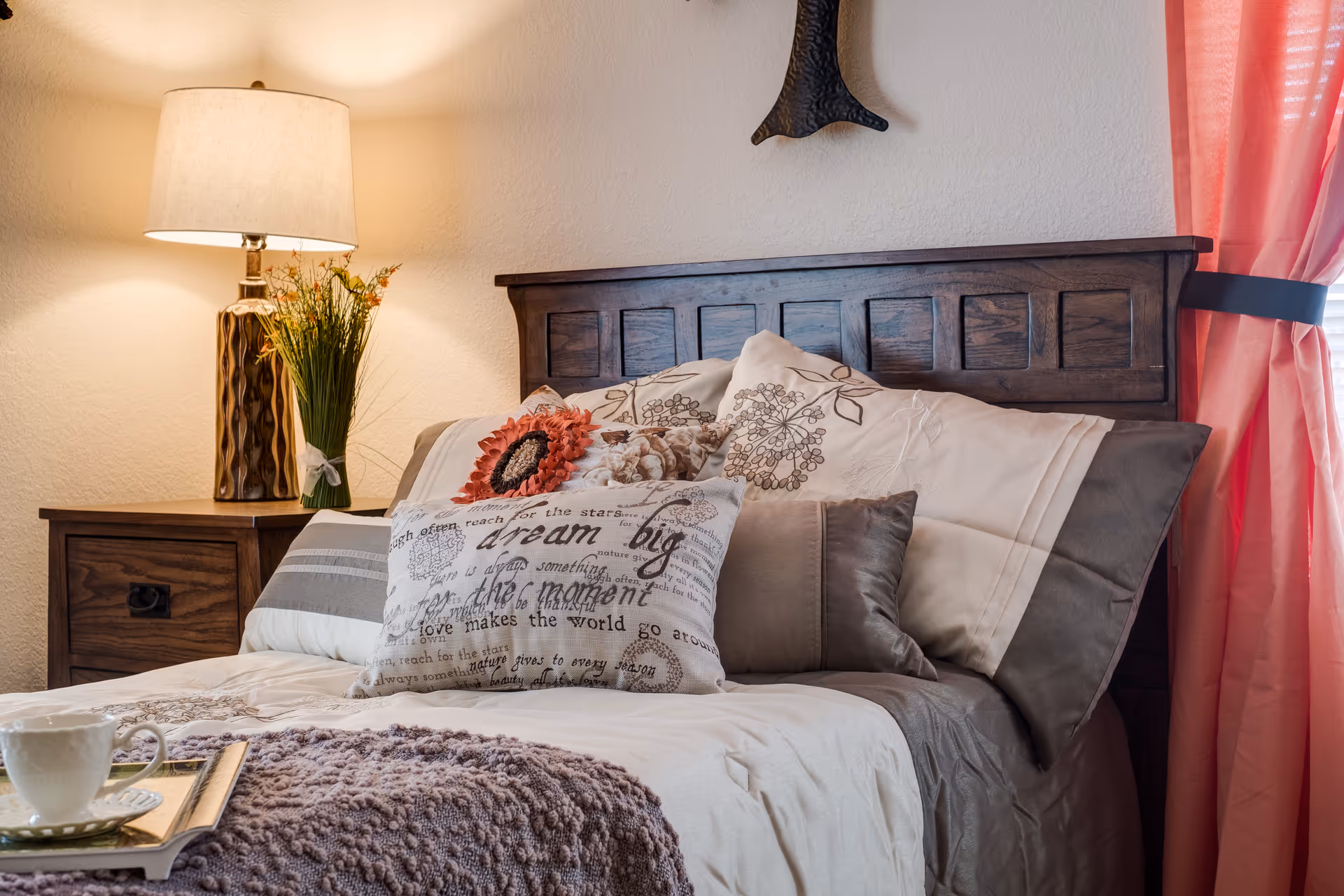 Sunlit bedroom with a wooden headboard, decorative pillows and throw on the bed, a lamp and vase of flowers on the nightstand, and pink curtains.