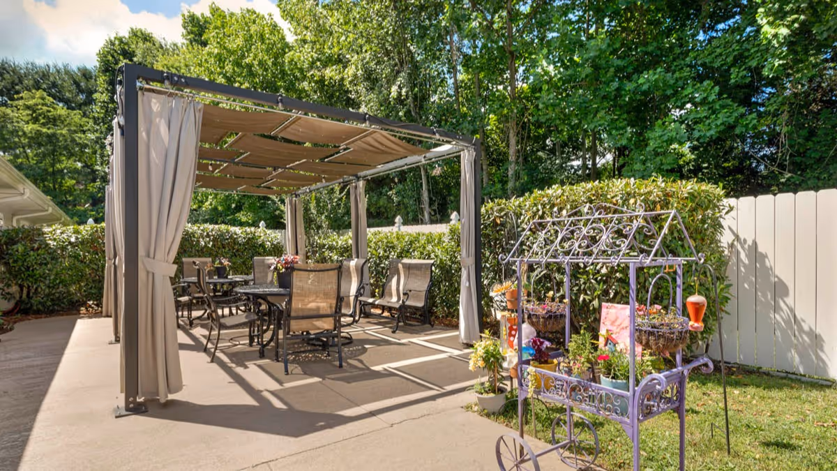 Outdoor patio area with a metal pergola covered by beige fabric curtains and roof panels, surrounded by green bushes and trees. Under the pergola are several black metal tables and chairs with beige mesh seats. To the right, there is a decorative purple metal plant stand with various potted flowers and plants. The area is paved with concrete and enclosed by a white fence.