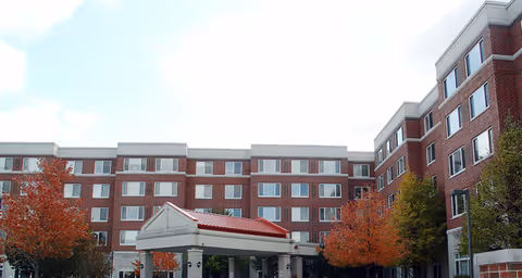 Exterior view of a multi-story brick senior living facility with a covered entrance and trees with autumn foliage in front.