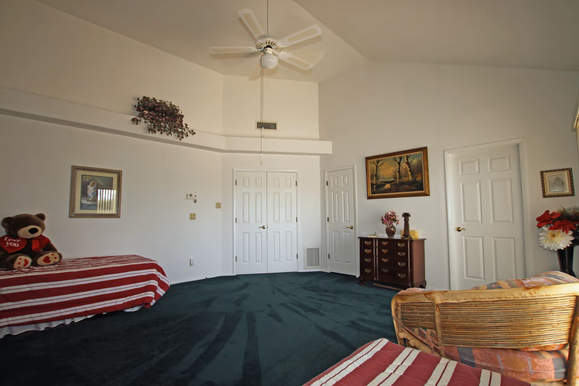 A spacious bedroom with dark green carpet, white walls, and a high ceiling with a ceiling fan. There are two beds with red and white striped bedding, a large teddy bear holding a heart that says 'Love you' on one bed, a wooden dresser with decorative items, framed paintings on the walls, and a wicker chair with a cushion. Three white doors are visible on the far wall.