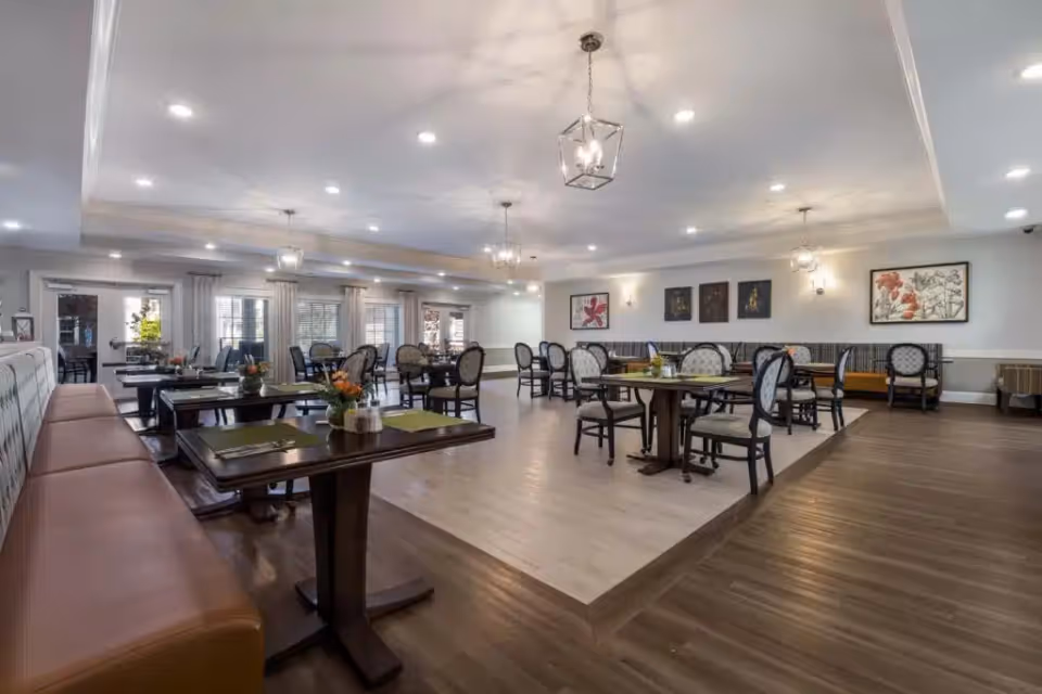 A spacious and well-lit dining room with multiple tables and chairs arranged neatly. The room features a combination of wooden and cushioned seating, pendant lights hanging from the ceiling, and framed artwork on the walls. Large windows with curtains allow natural light to enter the space.