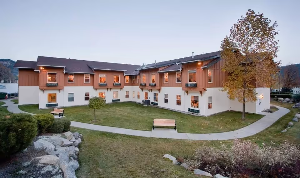 Exterior view of a two-story senior living facility building with a brown and white facade, surrounded by a well-maintained lawn, trees, bushes, and a paved walkway with benches.