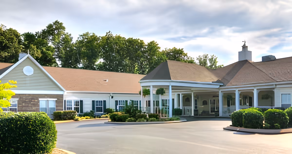Exterior view of a single-story senior living facility building with a covered entrance, surrounded by greenery and trees under a partly cloudy sky.