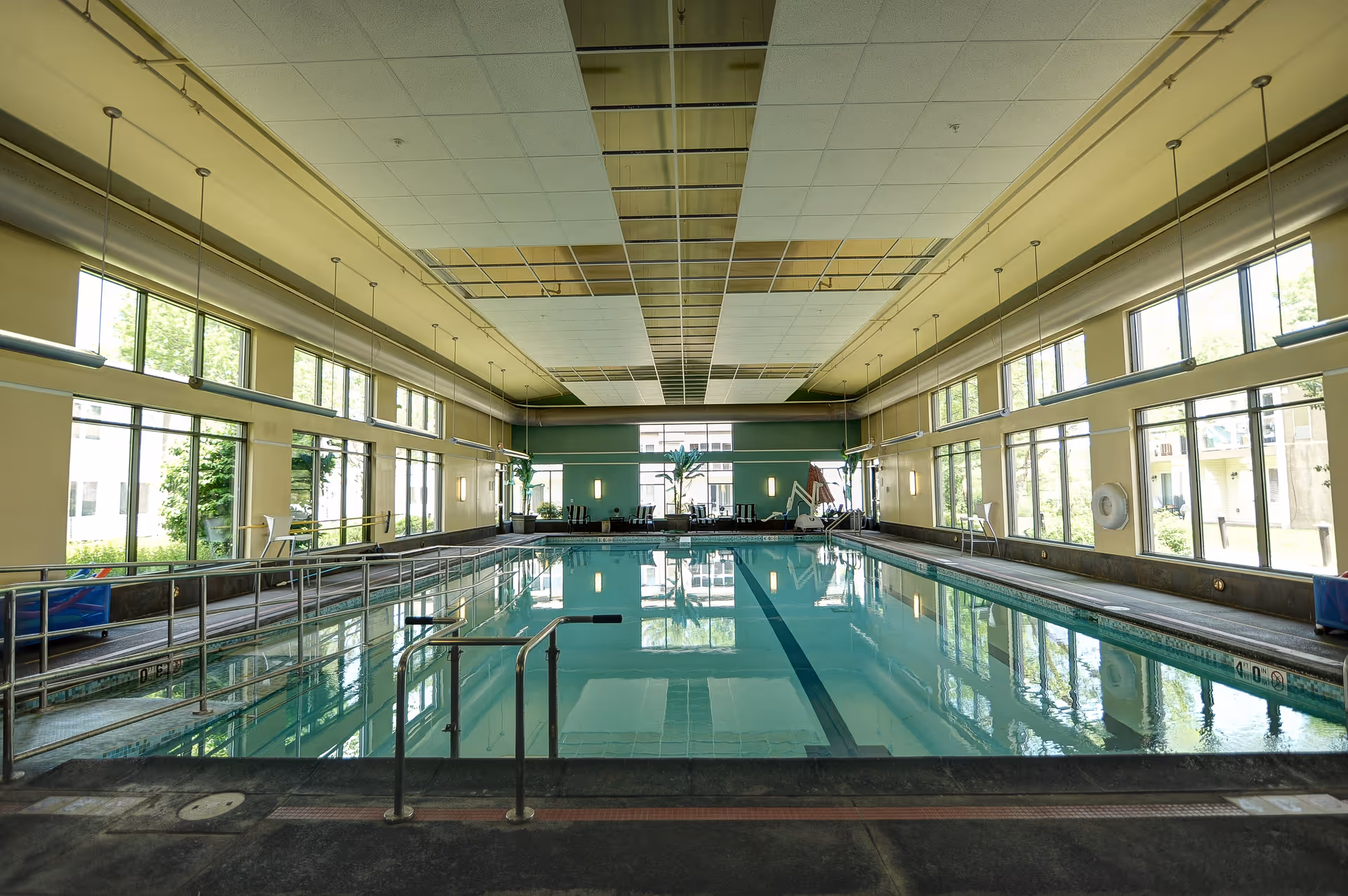 Indoor swimming pool with handrails and a lift for accessibility, surrounded by large windows letting in natural light. There are chairs and plants at the far end of the pool area.