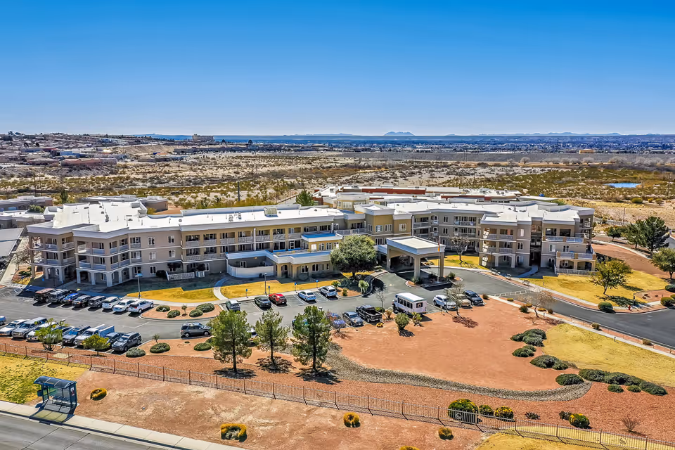 Aerial view of Solstice Senior Living at Las Cruces, a large multi-story senior living facility surrounded by parking lots, landscaped areas with trees and shrubs, and desert terrain in the background under a clear blue sky.