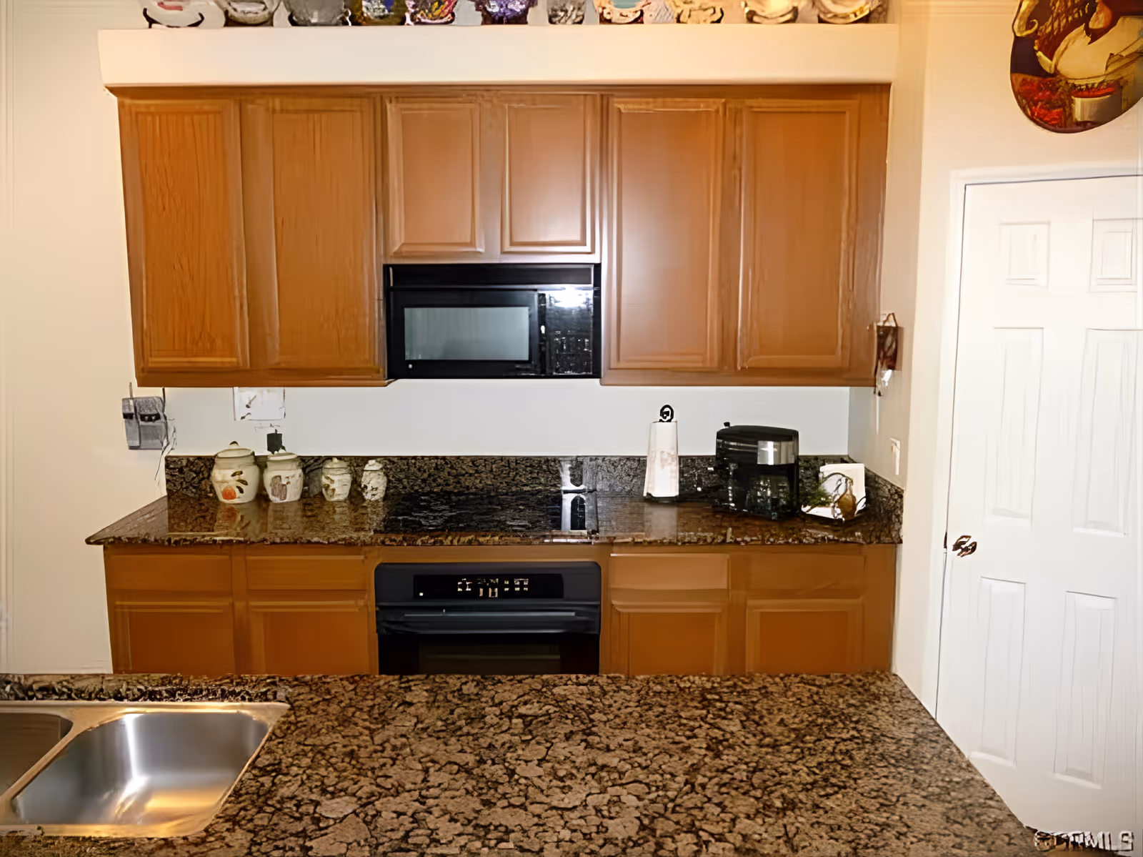 A kitchen with brown wooden cabinets, a black microwave oven, and a black stove. The countertop is made of dark granite with a speckled pattern and includes a double stainless steel sink. On the counter are several ceramic containers, a paper towel holder, and a small appliance. A white door is visible on the right side of the image.