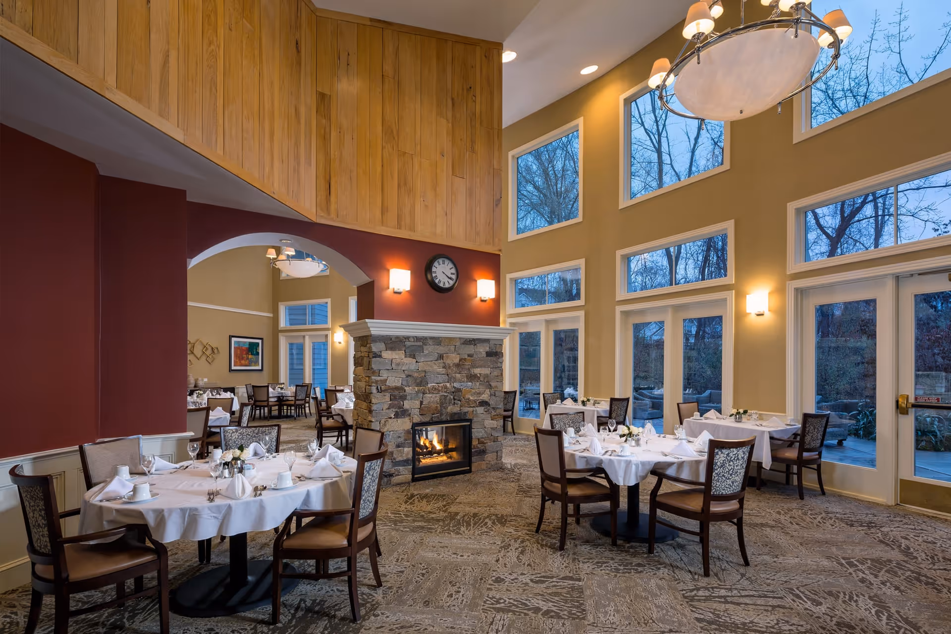 A spacious dining room with several round tables covered with white tablecloths and set with plates, cups, and napkins. The room features a stone fireplace in the center, large windows letting in natural light, and a chandelier hanging from the ceiling. The walls are painted in warm tones with wood paneling on the upper section and a clock above the fireplace.