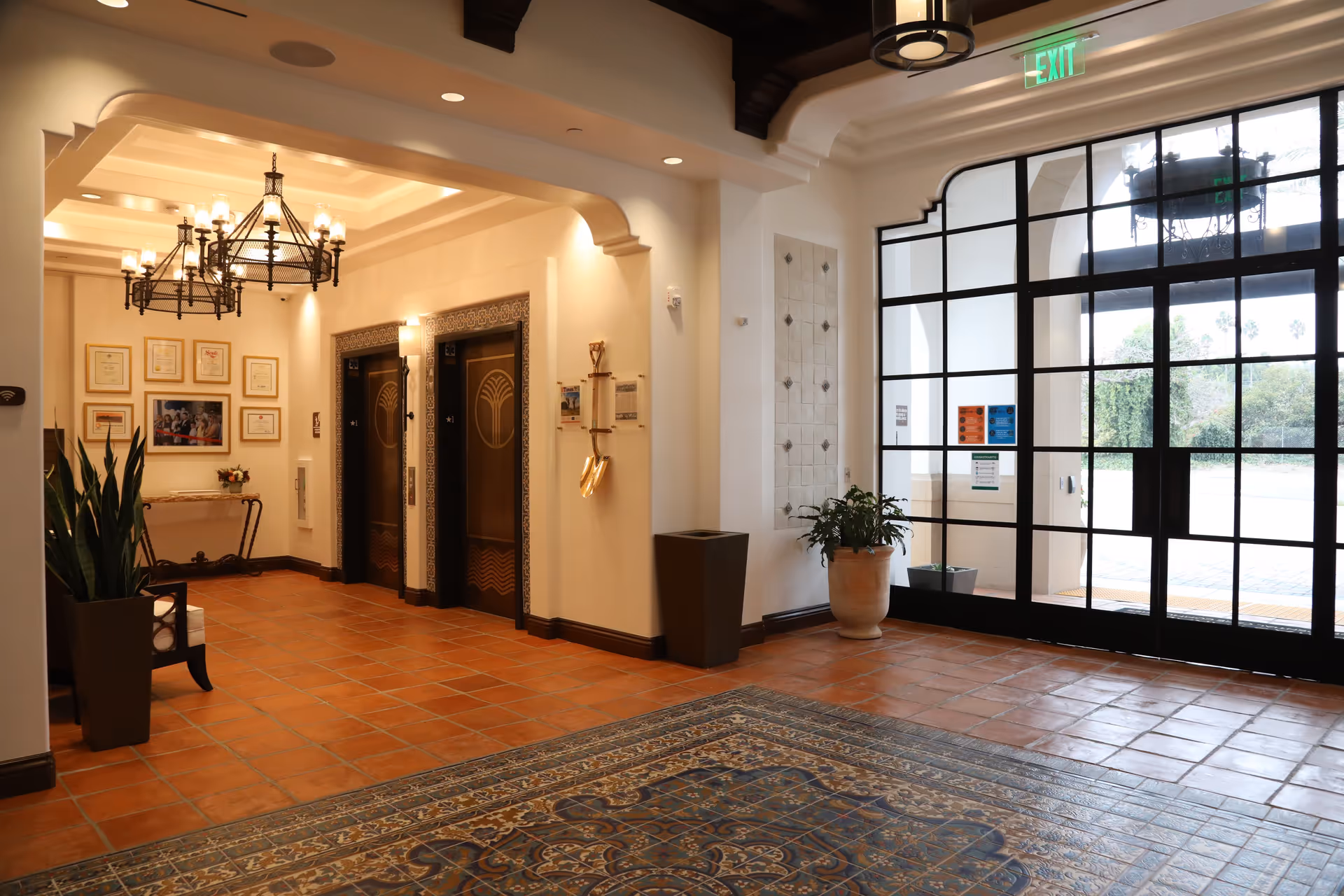 Interior view of a senior living facility lobby with terracotta tiled floor and a patterned rug. There are two elevators with decorative doors on the left wall, framed certificates and a photo above a small table, and potted plants near the elevators and entrance. The entrance features large glass doors with black frames letting in natural light. The ceiling has recessed lighting and a chandelier.