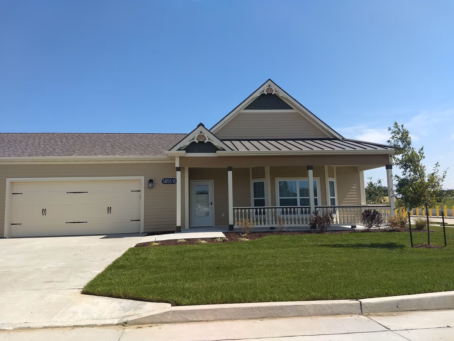 Single-story residential building with a beige exterior, a two-car garage, a covered front porch with decorative trim, and a well-maintained green lawn under a clear blue sky.