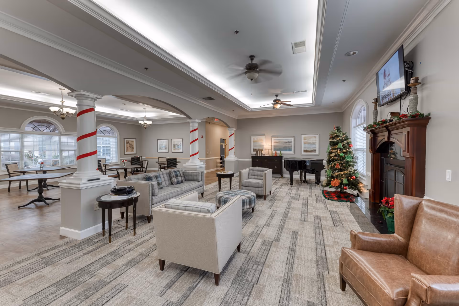A spacious and well-lit senior living facility common area decorated for Christmas with a Christmas tree, a fireplace with holiday decorations, a flat-screen TV mounted above the fireplace, a grand piano, and comfortable seating including sofas, armchairs, and ottomans. The room features columns wrapped with red ribbon, large windows with blinds, and a dining area with tables and chairs in the background.