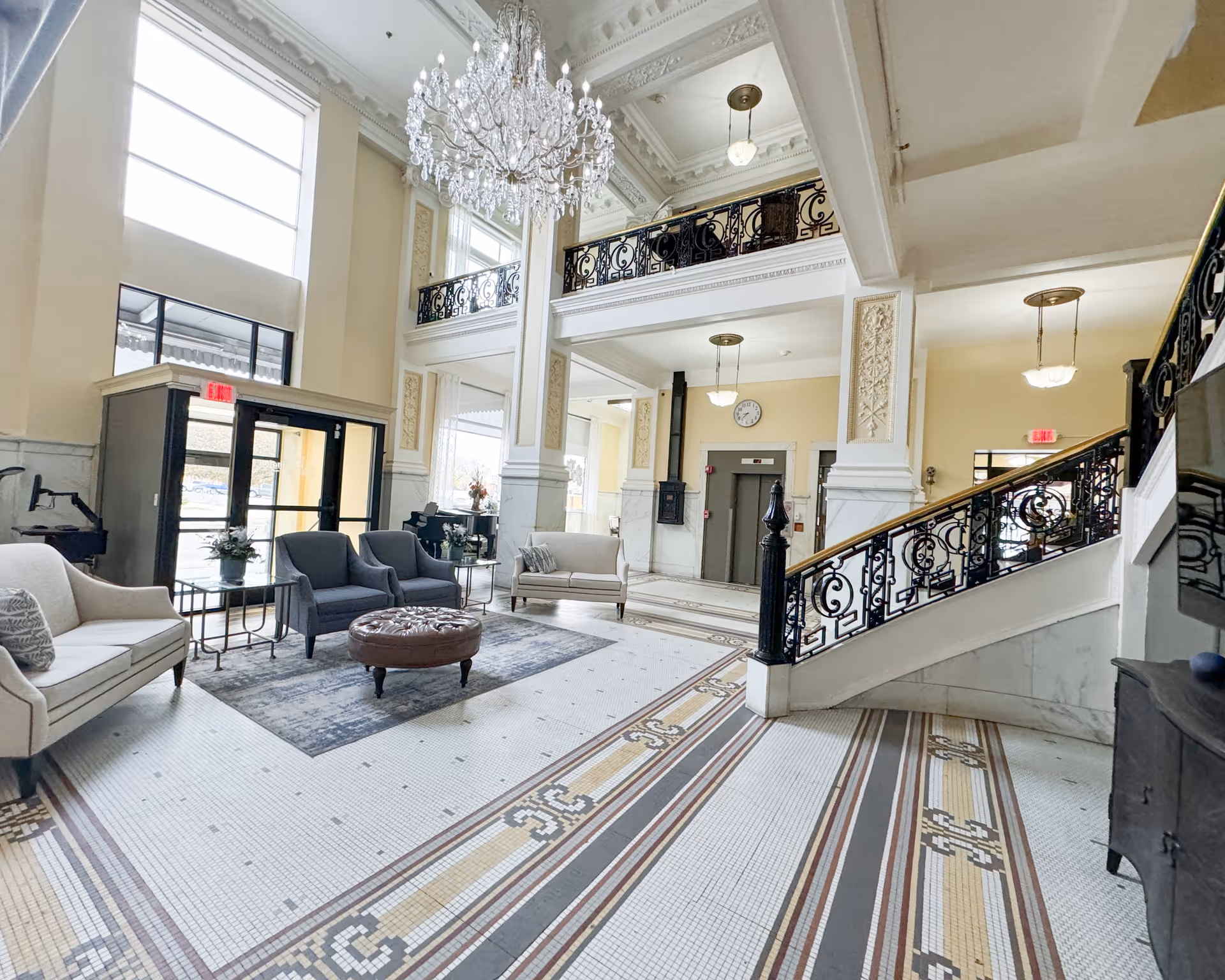 Spacious and elegant lobby area with high ceilings, a large crystal chandelier, mosaic tile flooring, and ornate railings on the staircase and balcony. The seating area includes sofas, armchairs, and a round ottoman on a rug near the entrance with large windows allowing natural light to fill the space.