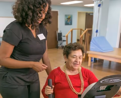 A caregiver in black scrubs assists an elderly woman wearing a red sweater and a gold necklace who is using a NuStep exercise machine in a well-lit room with wooden floors and medical equipment in the background.