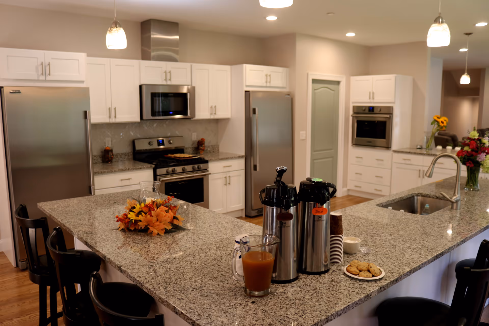 A modern kitchen with a large granite island, stainless steel appliances, coffee dispensers, and floral decorations.