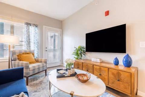 Bright living room with a patterned armchair, blue sofa, oval coffee table, wooden media console, and a wall-mounted TV near a glass door.